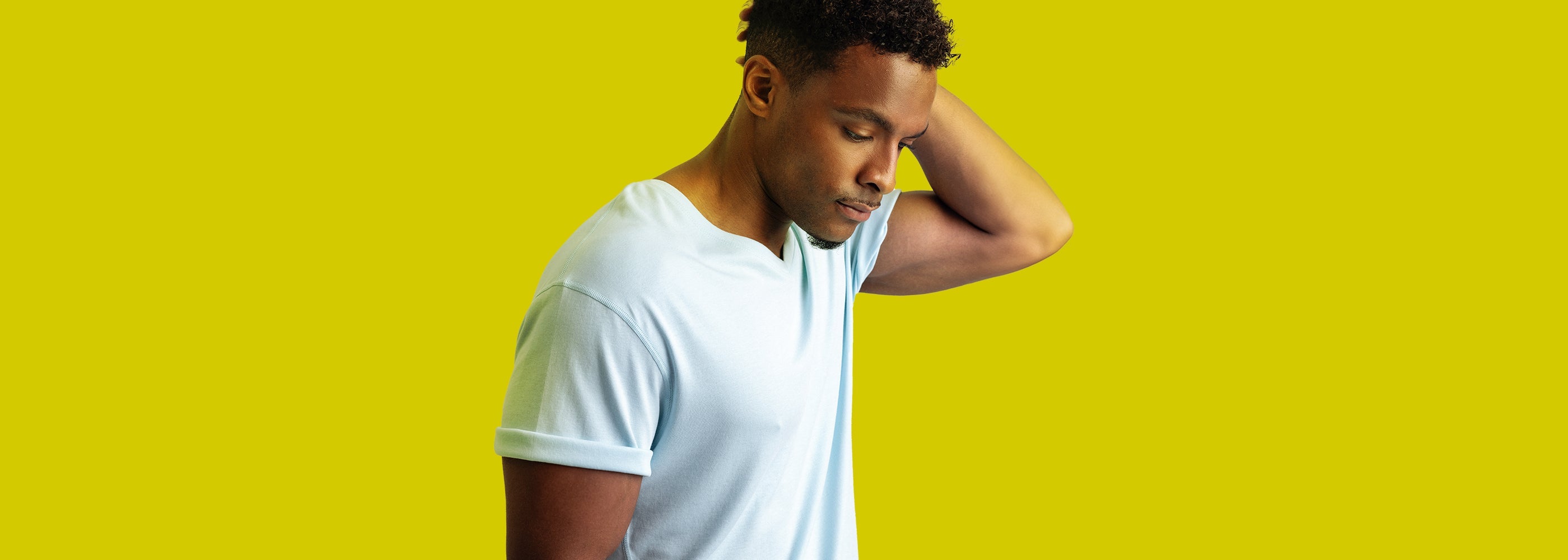 A young man wearing a light-colored T-shirt stands against a vibrant yellow background, with his head tilted down and one hand resting on the back of his neck. He has a thoughtful expression, and the soft fabric of the shirt highlights comfort and style. The overlay text reads, "50% sitewide offer."