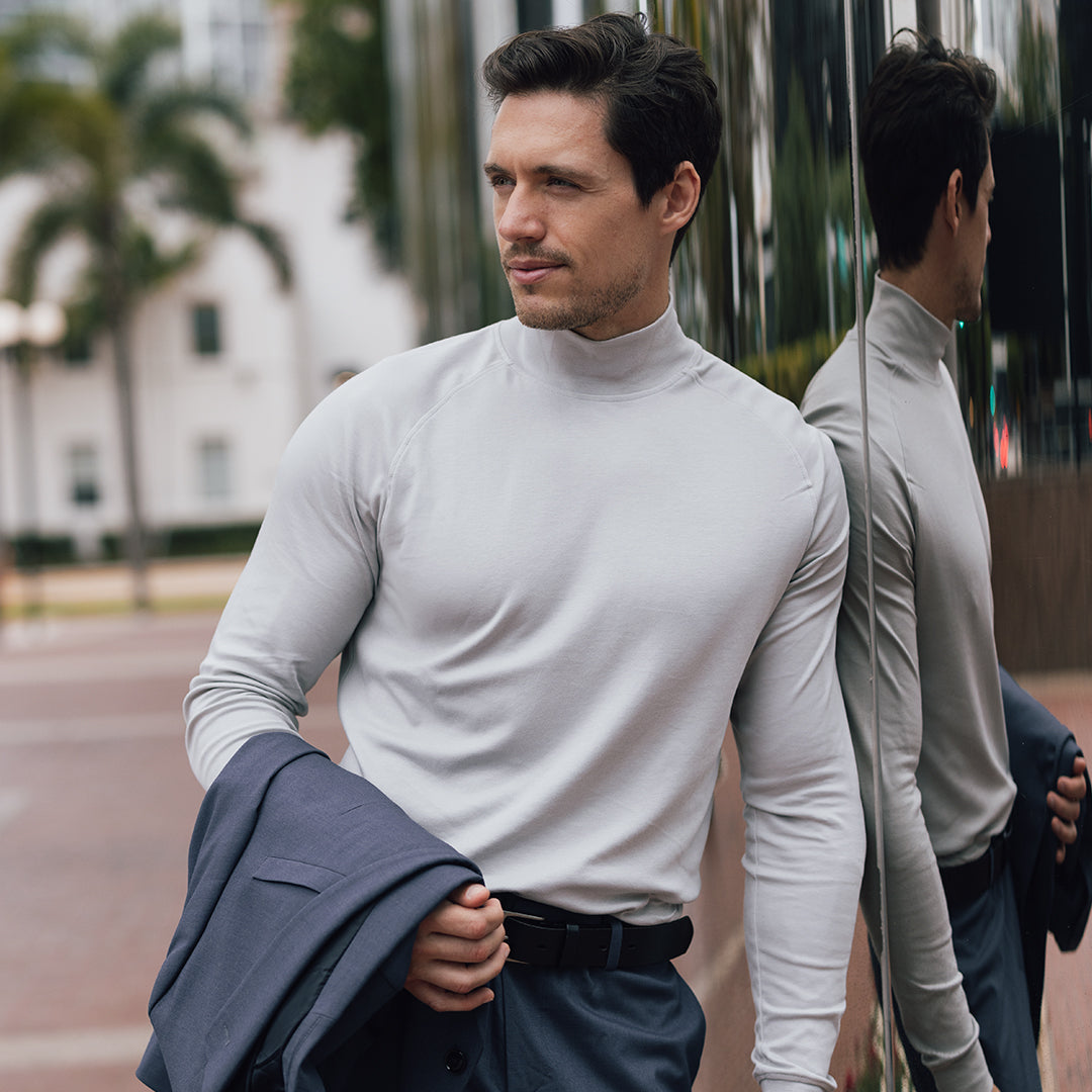 A man wearing a light grey Hi-Neck shirt stands confidently, leaning against a reflective surface outdoors. He holds a jacket casually draped over his arm, showcasing the Hi-Neck shirt's sleek design, which is perfect for layering under a jacket. The soft, fitted fabric enhances his sharp, stylish look. A white button with black text overlays the image, inviting users to "SHOP HI-NECK."